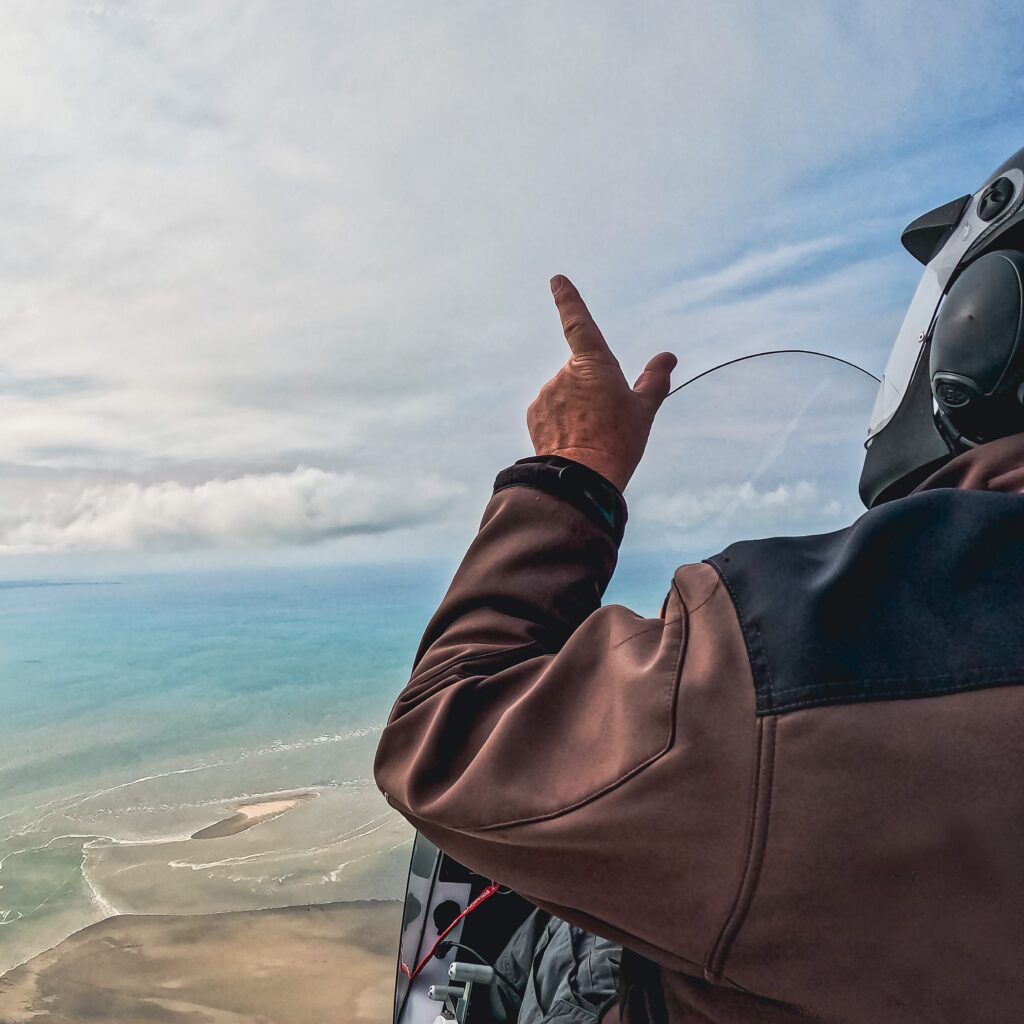 Instructeur en ULM désignant le ciel au-dessus du Mont Saint-Michel, vue aérienne sur la baie et l’océan.