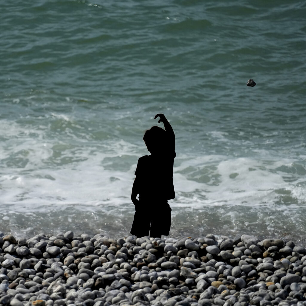 Enfant en ombre portée, bras levé, lance un caillou vers le ciel au-dessus de la mer, dessinant une trajectoire invisible.