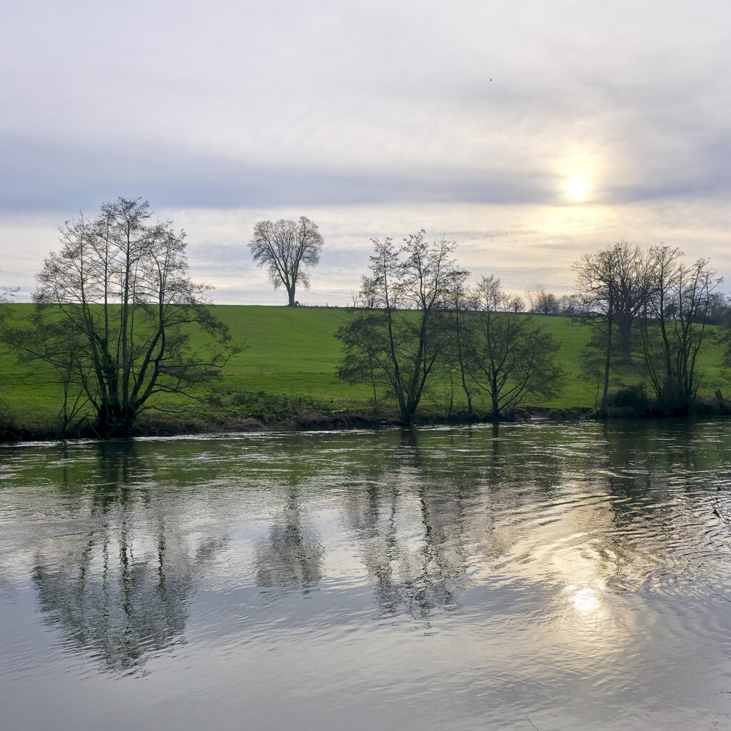 Paysage fluvial de l’Orne avec reflets des arbres sur l’eau calme et colline verdoyante.